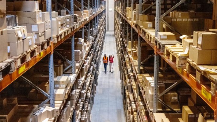 Large warehouse full of packages stacked high on shelves with two people walking down the center of the warehouse floow..