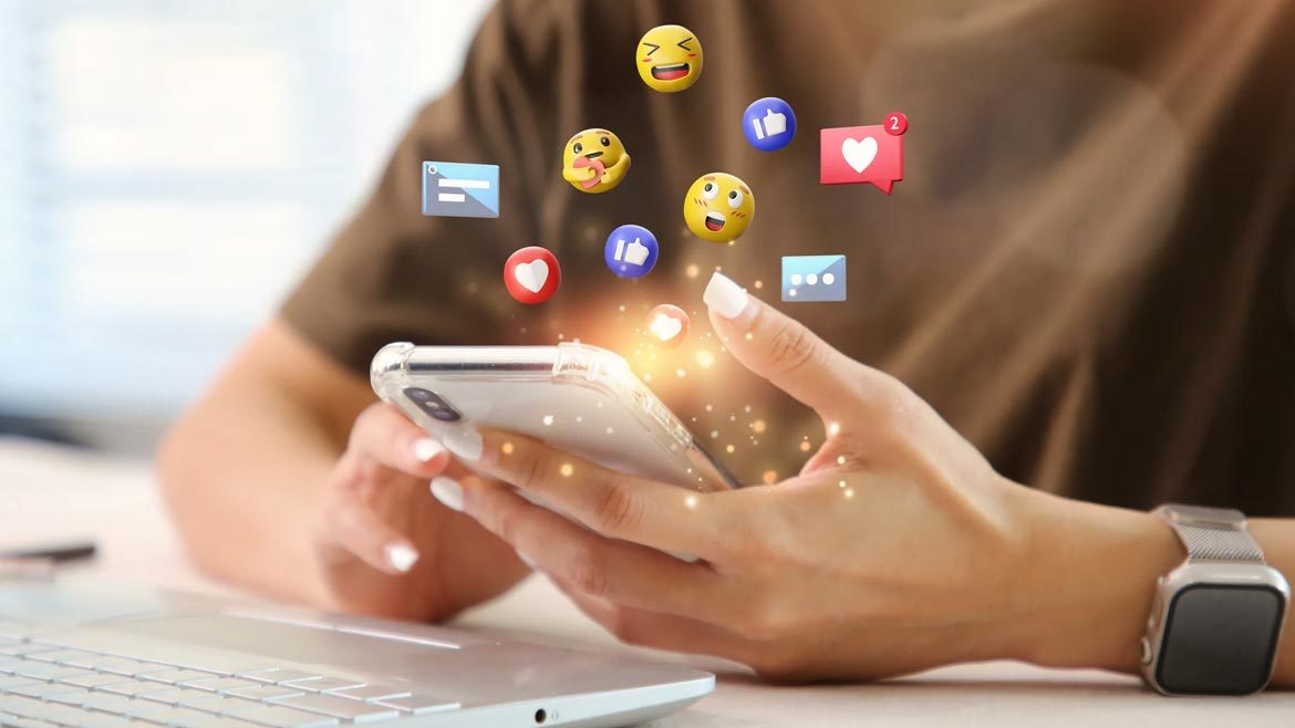 Woman using a laptop, on her mobile phone with icons and emojis floating above the phone