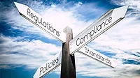 Signpost with words "Regulations", "Compliance", "Policies", and "Standards" pointing in different directions, with the cloudy skies above.