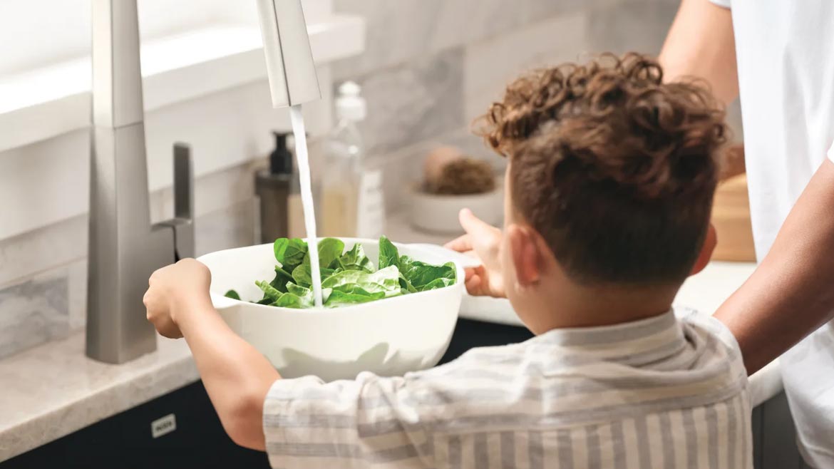 Little boy washing a bowl of salad in the sink.