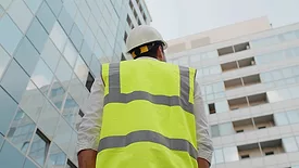 Low angle rear view of engineer with a white safety helmet and safety vest, looking up at modern building outdoors.