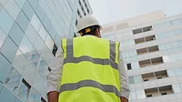 Low angle rear view of engineer looking up at modern building outdoors Low angle rear view of engineer with a white safety helmet and safety vest, looking up at modern building outdoors.
