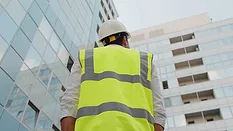 Low angle rear view of engineer with a white safety helmet and safety vest, looking up at modern building outdoors.