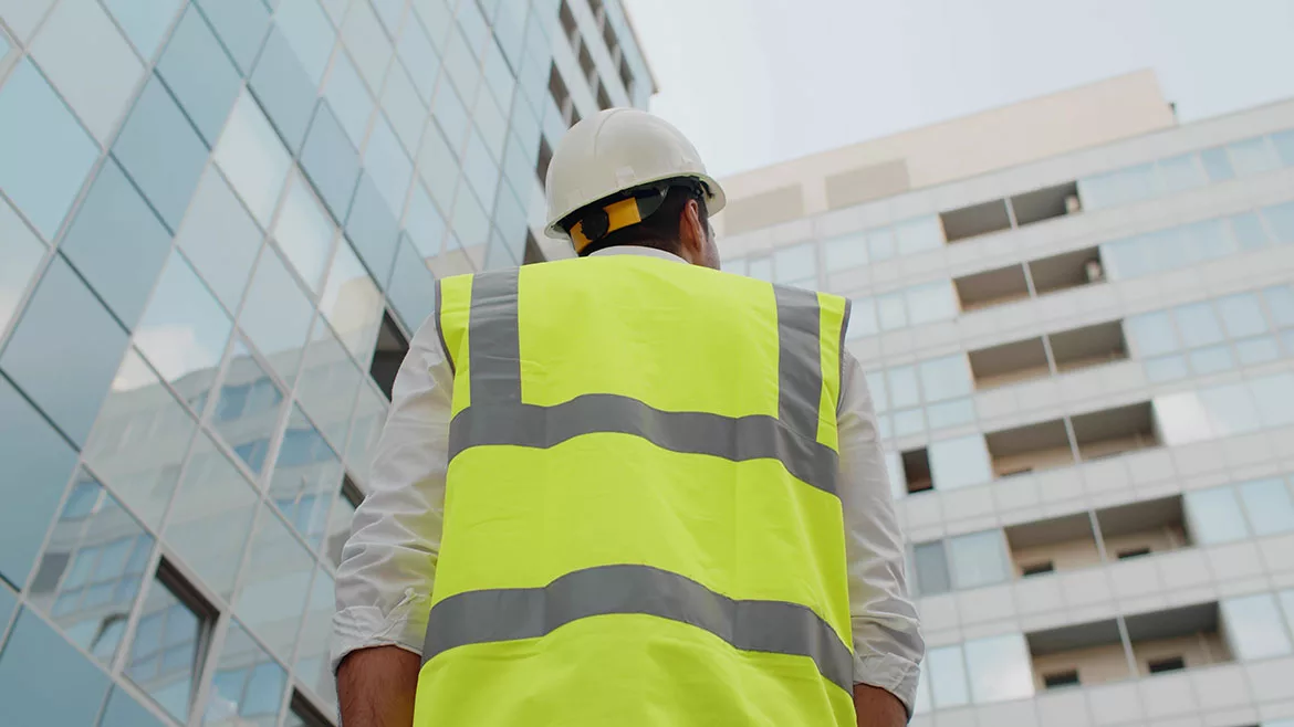 Low angle rear view of engineer with a white safety helmet and safety vest, looking up at modern building outdoors.