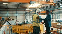 Two men, one in a safety vest and had and another in a business suit, shake hands in agreement while standing on the second level of a shipping warehouse.