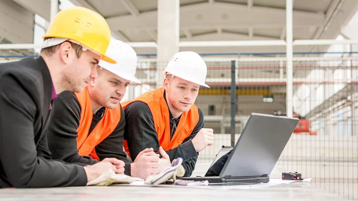 Three engineers checking plans on a laptop, the man on the left is wearing a yellow hard hat, the other two are wearing a white hard hat.