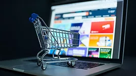 A miniature shopping cart on top of an open laptop, a shopping website blurred out and a black background.