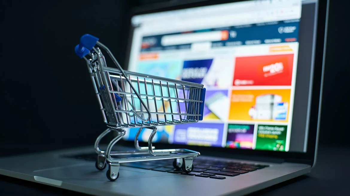 A miniature shopping cart on top of an open laptop, a shopping website blurred out and a black background.