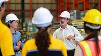 Manager talking with staff in warehouse, all wearing hard hats. A woman holing a tablet is facing the manager, while three people's backs are facing the front.