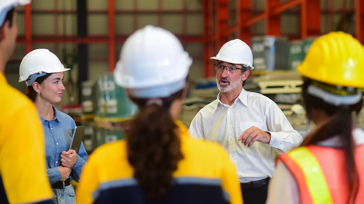 Manager talking with staff in warehouse, all wearing hard hats. A woman holing a tablet is facing the manager, while three people's backs are facing the front.