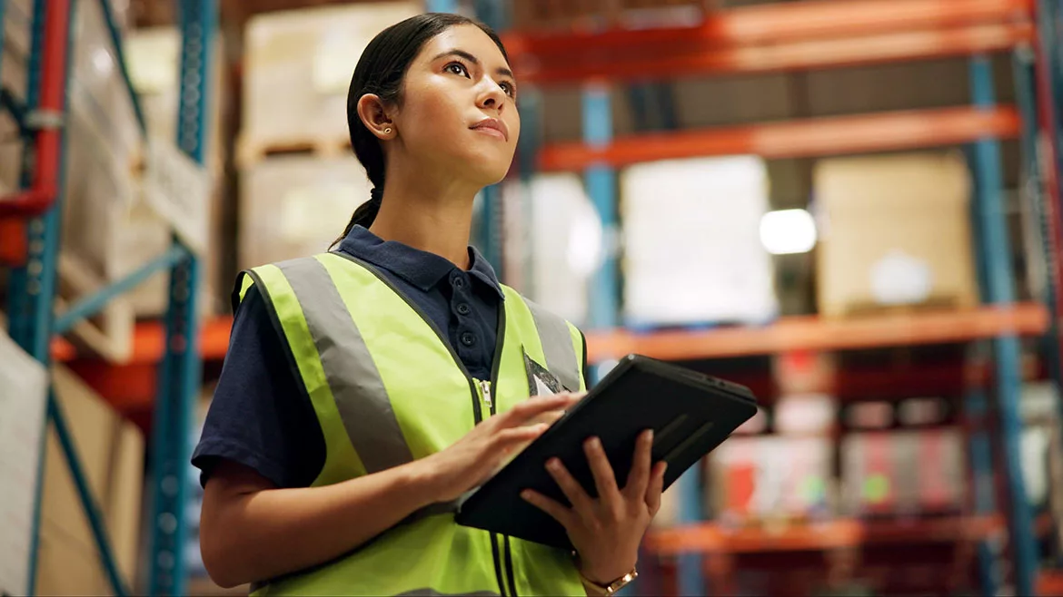 Female worker in safety vest is holding a laptop while working in a warehouse.