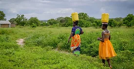Two women walking across a grassy field. They're carrying water jugs on their head.