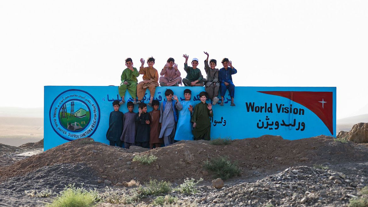 Young boys posing in front of a World Vision sign in Afghanistan. Six are sitting on top of the sign and seven standing and leaning against the sign.