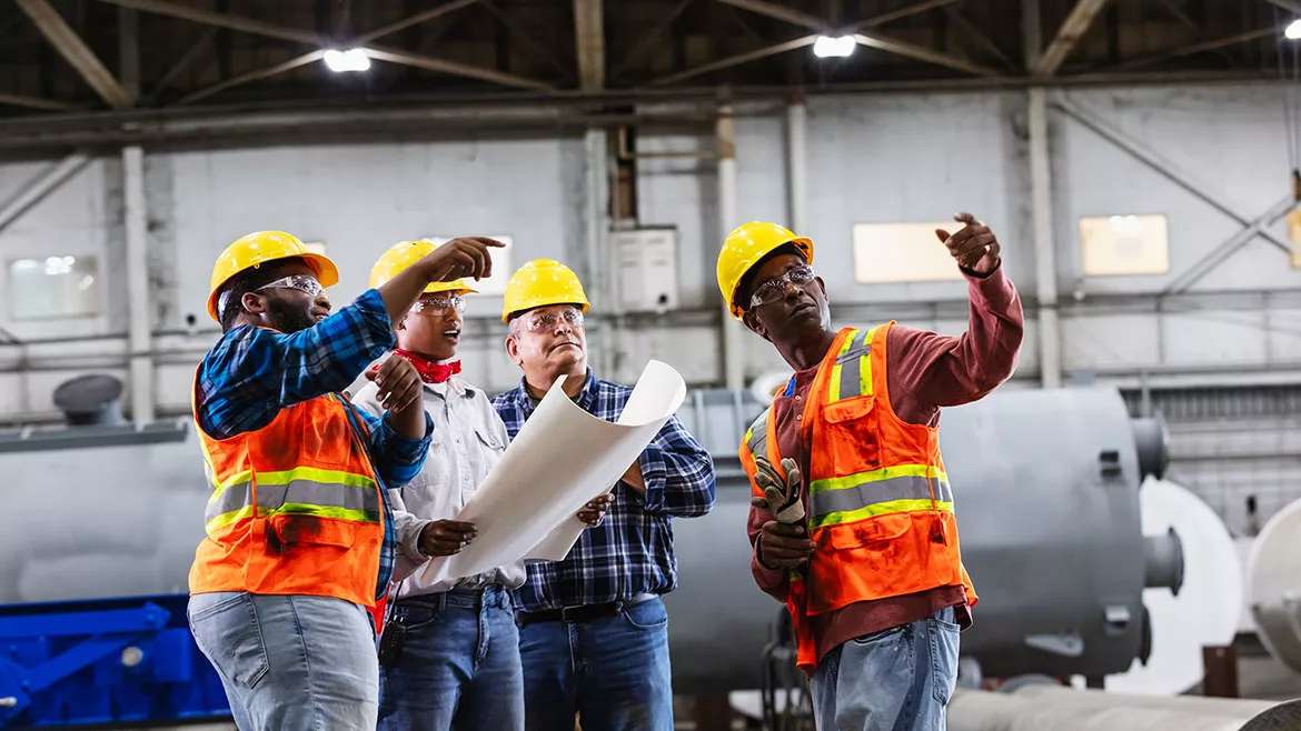 A diverse group of four engineers working in a factory is having a meeting. The female engineer is holding a set of blueprints. They are all looking up at something, two of them pointing.