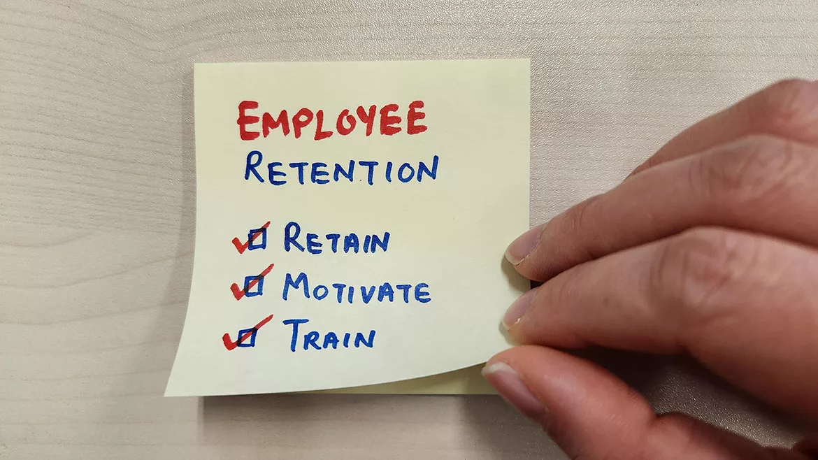 A sticky note on a wall with writing: EMPLOYEE RETENTION, three words each with a checked box - Retain, Motivate, Train