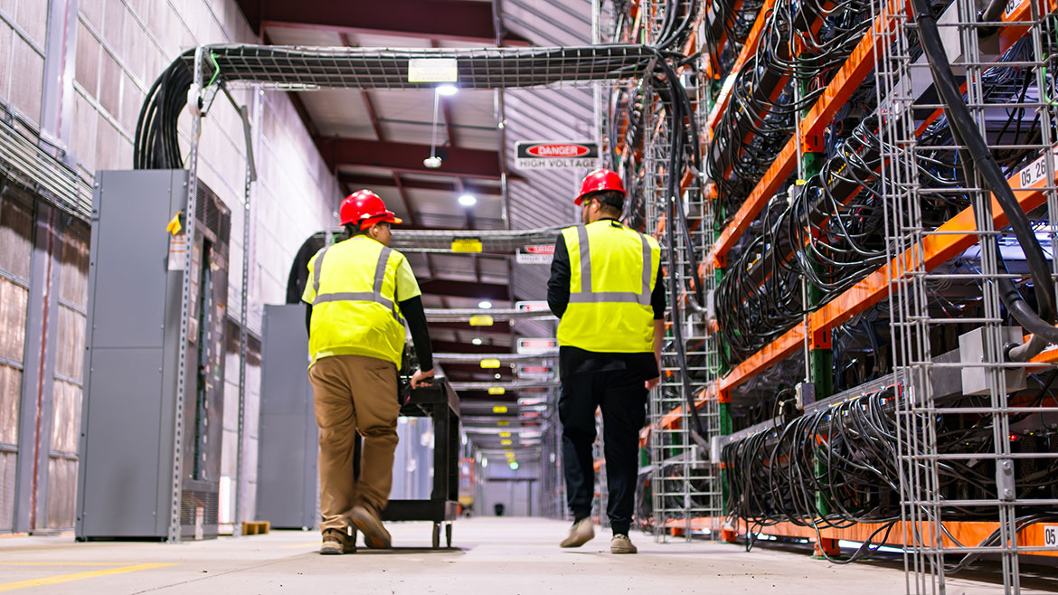 Two technicians wearing PPE walking past racks of equipment in a data center for cryptocurrency mining, cloud services and AI computing in a large, temperature controlled warehouse in a remote location in Stutsman County, North Dakota.