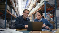 Two managers checking for delivery order, stock or ecommerce items on a laptop, with tall warehouse shelves behind them.