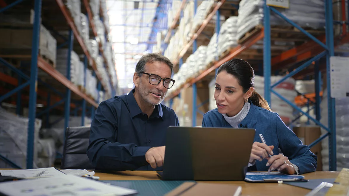 Two managers checking for delivery order, stock or ecommerce items on a laptop, with tall warehouse shelves behind them.