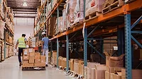 Two warehouse workers are moving boxes in a large distribution center.