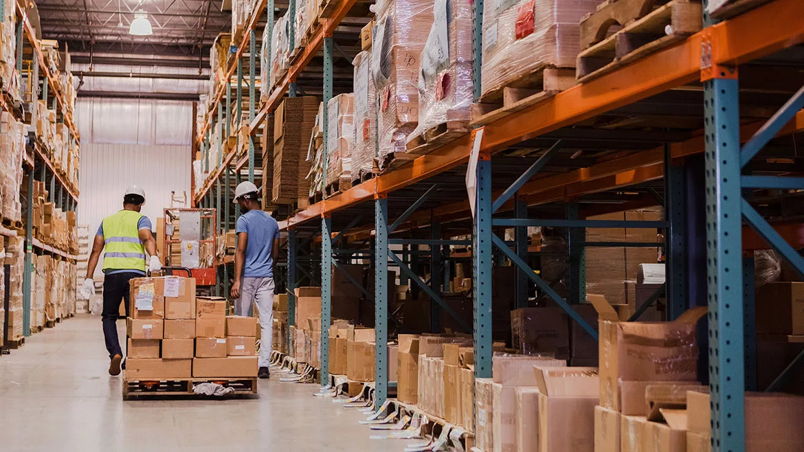 Two warehouse workers are moving boxes in a large distribution center.