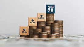 New Year concept wooden blocks with 'GOAL', 'PLAN', 'ACTION', AND '2026' text, checkmarks, and a bullseye icon. The blocks are stacked on top of coins, which are stacked on top of paper cash.