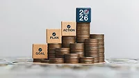 New Year 2026 concept wooden blocks with text on top of stacked coins New Year concept wooden blocks with 'GOAL', 'PLAN', 'ACTION', AND '2026' text, checkmarks, and a bullseye icon. The blocks are stacked on top of coins, which are stacked on top of paper cash.