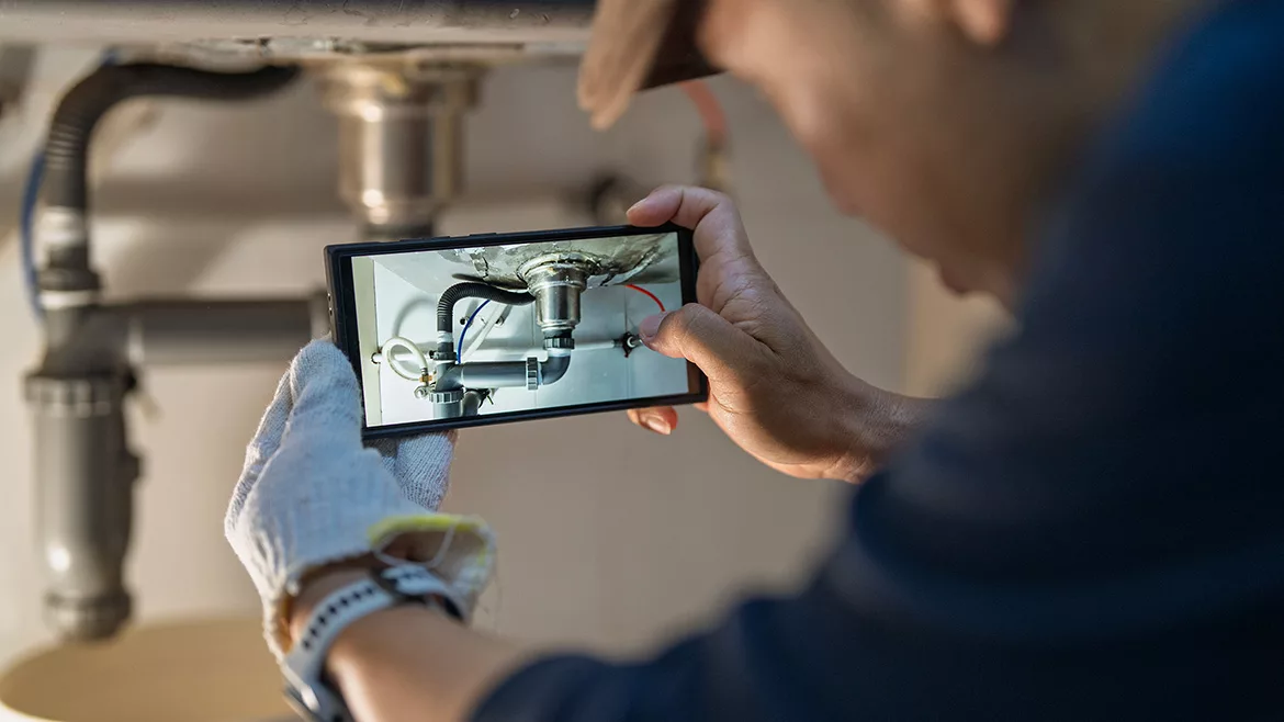 Man using a smartphone to document a leaking kitchen sink pipe at home.