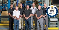 Merit Brass Co.group photo in a warehouse with a conveyor belt, stairs and industrial equipment in the background.