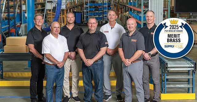 Merit Brass Co.group photo in a warehouse with a conveyor belt, stairs and industrial equipment in the background.