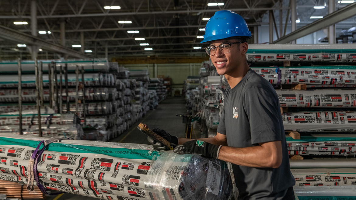 Smiling worker in hard hat handles wrapped stainless steel pipes in a warehouse, more wrapped pipes on shelves in the background.