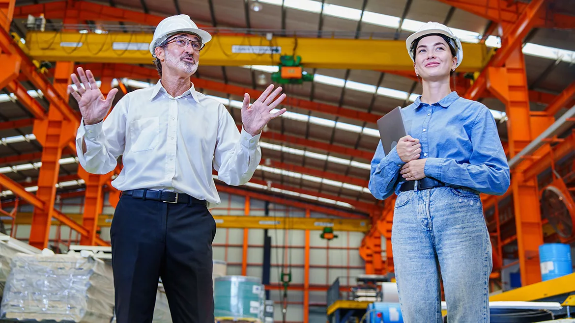 Two engineers wearing white hardhats inspect a factory, the man making a gesture, the woman holding a tablet.